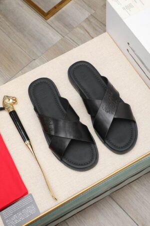 Top-down view of black Berluti Scritto Venezia leather cross-strap sandals with black contoured soles displayed on a beige surface next to decorative accessories.