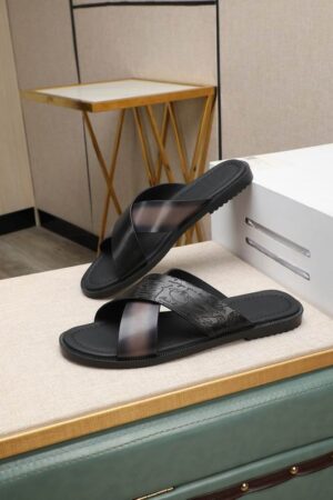 Top-down view of Berluti Scritto Venezia leather cross-strap sandals in a nero grigio patina with black contoured soles, displayed on a beige textured surface next to a red cloth and decorative accessories.