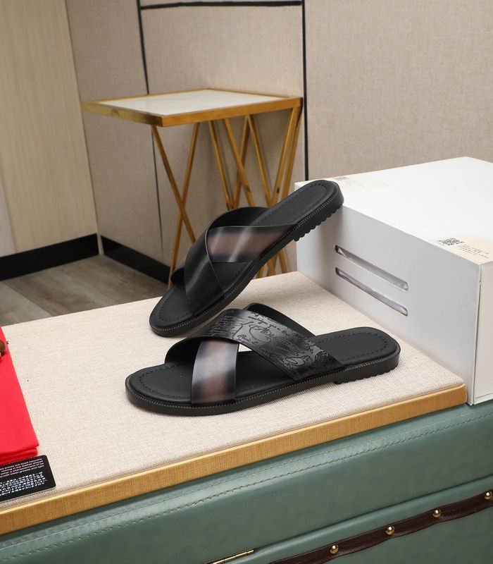 Top-down view of Berluti Scritto Venezia leather cross-strap sandals in a nero grigio patina with black contoured soles, displayed on a beige textured surface next to a red cloth and decorative accessories.