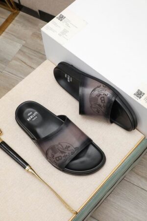 Top-down view of Berluti Scritto Venezia leather slides in a nero grigio patina with black contoured soles, displayed on a beige surface next to a red cloth and decorative accessories.
