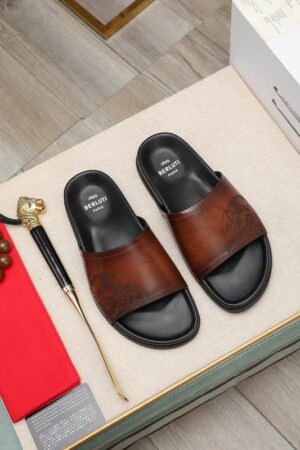 Top-down view of Berluti Scritto Venezia leather slides with a brown and black patina and black contoured soles, displayed on a beige surface with accessories.