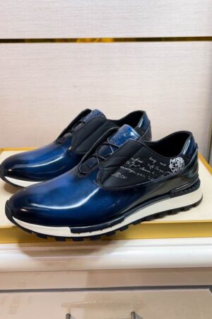 Top-down view of Berluti Fast Track sneakers in a glossy blue patina leather with Scritto engravings and black laces on a white shoebox.