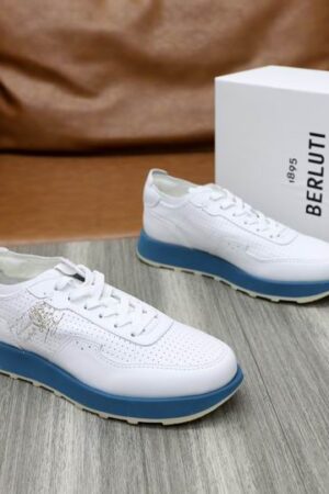 Top-down view of white Berluti Playtime sneakers featuring Scritto leather engravings and a bold blue midsole on a wooden surface.