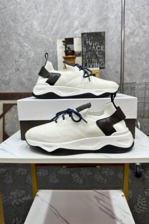 Pair of luxury Berluti Shadow sneakers in white knit fabric with brown leather heel accents and navy blue laces on a marble surface.