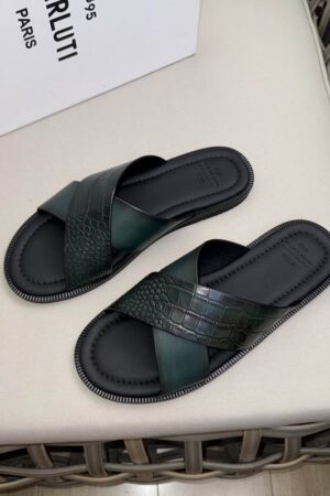 Top-down view of green Berluti Venezia and alligator-embossed leather cross-strap sandals with black contoured soles displayed on a beige surface.