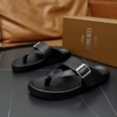 Pair of black Berluti Scritto leather thong sandals with silver buckles and black contoured soles displayed on a black table with a brown box in the background.