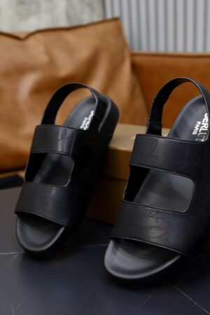 Top-down view of black Berluti Lorenzo leather sandals featuring iconic Scritto engravings and ergonomic black footbeds on a dark marble table.