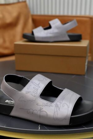 Top-down view of grey Berluti Lorenzo leather sandals featuring Scritto engravings and black ergonomic footbeds on a dark marble table.