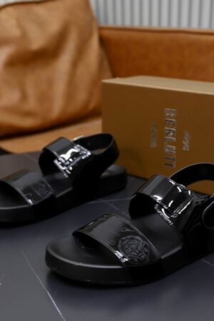 Side view of glossy black Berluti Lorenzo patent leather sandals with Scritto engravings, silver buckles, and thick black comfort soles on a dark table.