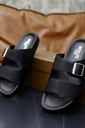 Pair of black Berluti Scritto leather double strap sandals with a silver buckle and black contoured soles displayed on a black table with a brown box.
