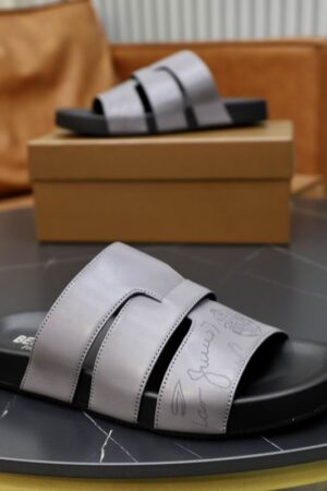 Pair of grey Berluti Scritto leather sandals with a unique H-strap design and black contoured soles displayed on a black table with a brown box.