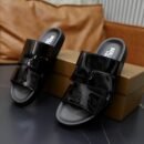 Top-down view of black patent leather Berluti Scritto sandals with a unique H-strap design and black contoured soles displayed on a grey table.