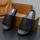 Top-down view of black Berluti Scritto leather slides with a detailed crest engraving and black contoured soles displayed on a grey table.
