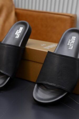 Top-down view of black Berluti Scritto leather slides with a detailed crest engraving and black contoured soles displayed on a grey table.