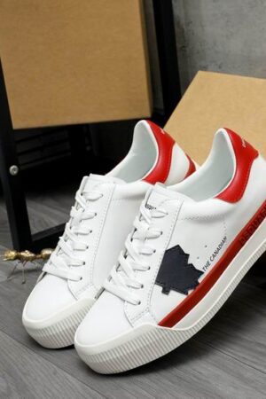 A pair of DSQUARED2 The Canadian low-top sneakers in white leather, featuring a red heel tab, a red side stripe, and a navy blue maple leaf patch, leaning against a cardboard box.
