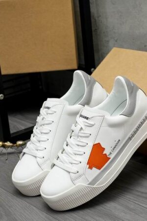 A pair of DSQUARED2 The Canadian low-top sneakers in white leather, featuring a matte gray heel tab, a gray side accent stripe, and a bright orange maple leaf patch, leaning against a brown cardboard box.