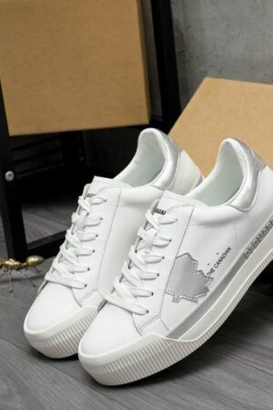 A pair of DSQUARED2 The Canadian low-top sneakers in white leather, featuring a metallic silver heel tab, a silver side stripe, and a silver maple leaf patch, leaning against a cardboard box.