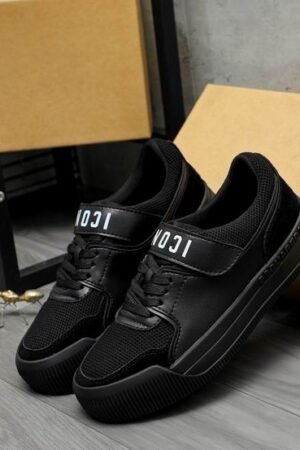 A pair of DSQUARED2 Icon low-top sneakers in all-black leather and mesh, featuring a prominent midfoot strap with white "ICON" branding, leaning against a cardboard box.