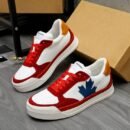 A pair of DSQUARED2 The Canadian low-top sneakers featuring a white leather upper, red suede panels, a blue maple leaf logo, and a yellow heel tab, leaning against a box.
