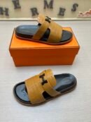 A pair of Hermès Chypre sandals in Mustard Yellow crocodile-embossed leather with black soles and matte black "H" hardware, resting on an orange designer box.