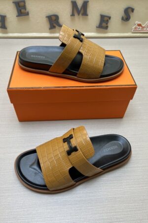A pair of Hermès Chypre sandals in Mustard Yellow crocodile-embossed leather with black soles and matte black "H" hardware, resting on an orange designer box.