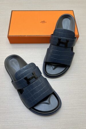 A pair of Navy Blue Hermès Chypre sandals with crocodile-embossed leather straps and black anatomical footbeds, displayed with a classic orange designer box.