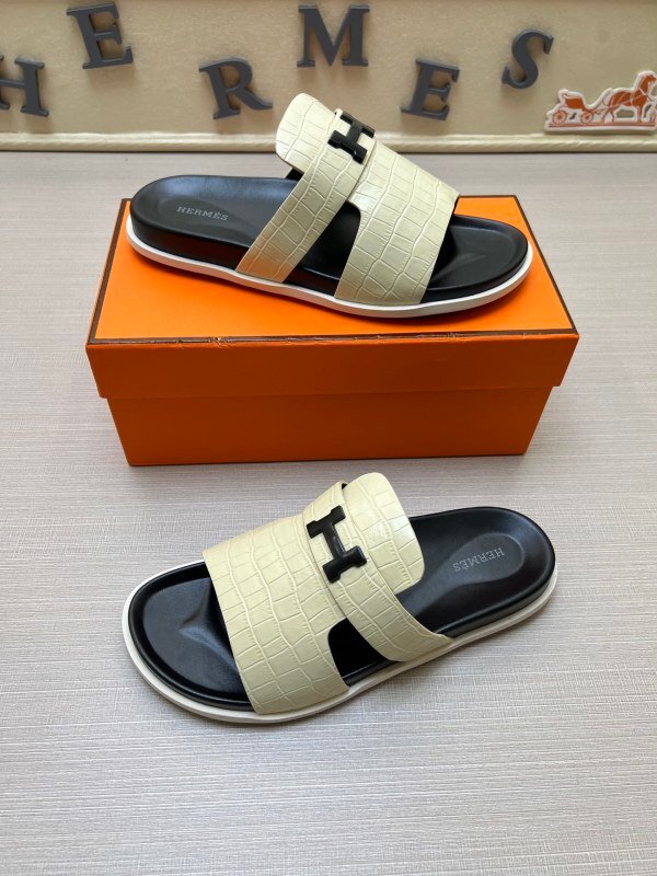 Top-down view of Hermès Chypre sandals in Chalk White crocodile-embossed leather featuring black footbeds and matte black hardware, placed in front of an orange luxury box.