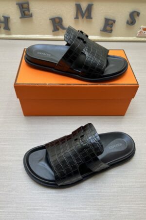 A pair of all-black Hermès Chypre sandals with crocodile-embossed leather straps and matching black soles, positioned in front of a signature orange designer box.