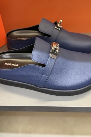Pair of luxury Hermès Oz mules in navy blue Epsom leather featuring the silver Kelly buckle on a neutral boutique shelf.