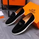 Pair of luxury black suede loafers with silver H buckles and white rubber soles displayed against orange designer boxes.