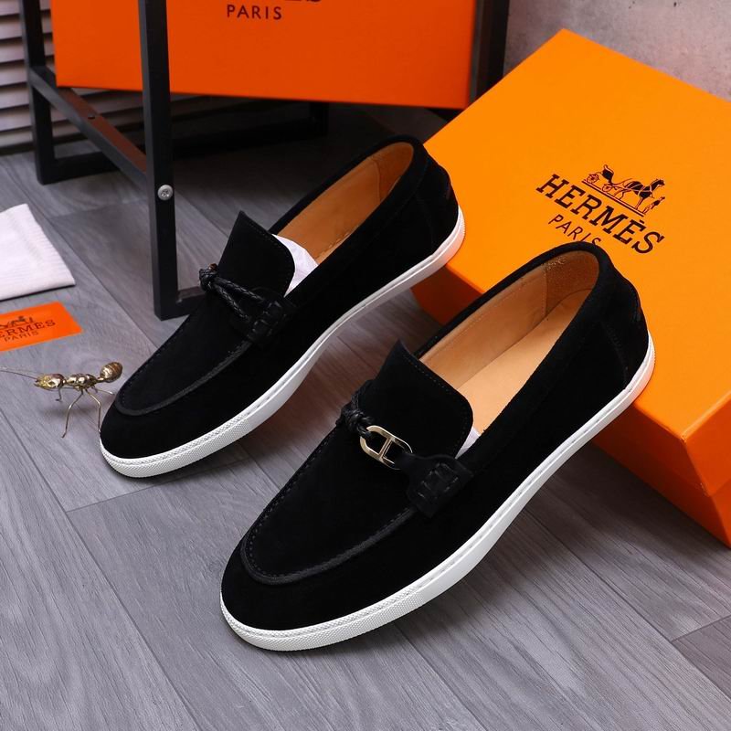 Pair of luxury black suede loafers with silver H buckles and white rubber soles displayed against orange designer boxes.