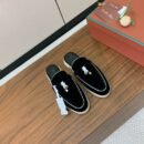 A pair of premium black suede backless mule loafers with white piping and silver charms, displayed on a wooden surface next to an orange shoe box.