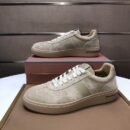 A side profile view of a pair of men's premium sand beige suede sneakers with cream laces and a matching taupe rubber sole, stacked on an orange luxury shoe box.