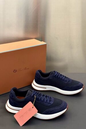 A pair of premium deep navy blue Loro Piana low-top sneakers with mesh and suede panels and a thick white sole, resting on a dark gray surface next to a brand box.