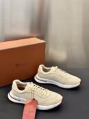 A pair of premium men's beige knit and suede sneakers with white rubber soles displayed on a dark surface next to an orange luxury shoe box.