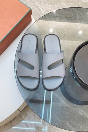 A top-down view of a pair of premium men's gray leather slide sandals with asymmetric cutout straps, displayed on a glass table next to an orange shoe box.