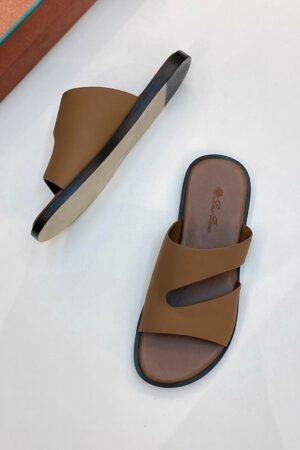 A top-down view of a pair of premium men's tan leather slide sandals with asymmetric cutout straps, displayed on a clean white surface next to an orange shoe box.