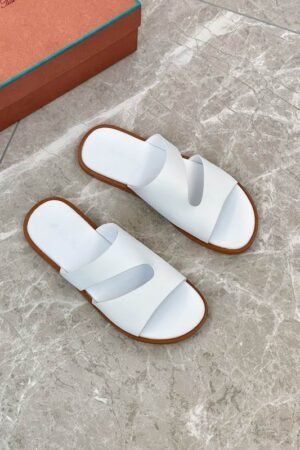 A top-down view of a pair of men's premium pure white leather slide sandals with asymmetric cutout straps and a brown leather trim, displayed on a glass table.