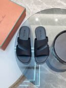 A top-down view of a pair of men's premium matte black leather slide sandals with asymmetric cutout straps, displayed on a grey marbled surface next to an orange shoe box.